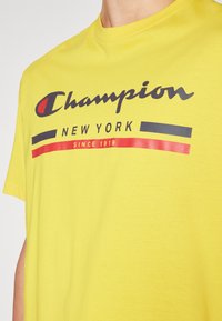 Camiseta de algodón amarilla con mangas cortas, que presenta el texto y elementos gráficos en negro y rojo "Champion New York".