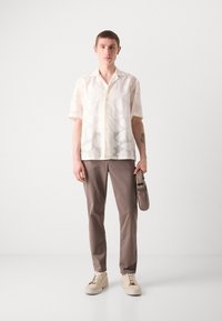 Korte mouwen, lichtgepatroneerd shirt van doorschijnende off-white stof, gecombineerd met een bruine broek en crèmekleurige sneakers, met een kleine grijze tas.