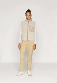 Μπεζ γιλέκο φλις με κλείσιμο φερμουάρ, τσέπη μπροστά στο στήθος και αντίθετες καφέ λεπτομέρειες, φορεμένο πάνω από μακριά μανίκια πουκάμισο και μπεζ παντελόνι.