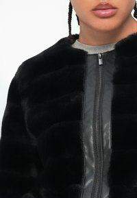 Crna umjetna krznena jakna na zipper s kožnim detaljima, koja ima teksturiranu završnu obradu i rebrasti dizajn duž tijela za dodatne detalje.