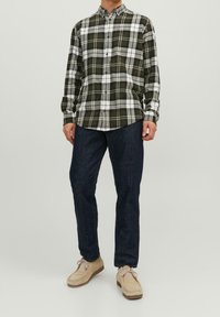 Camicia a quadri verde e bianca con bottoni e maniche lunghe, abbinata a pantaloni di jeans blu scuro e scarpe beige chiaro.