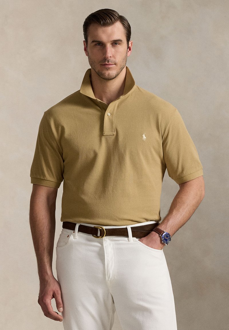 Tan polo shirt with a collar, short sleeves, and a small embroidered logo. Worn with white pants and a brown belt, featuring a blue watch.