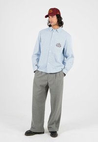 Blau-weiß gestreiftes Hemd mit Knöpfen und besticktem Detail, graue Faltenhose und eine kastanienbraune Mütze mit goldener Schrift. Schwarze Schuhe runden das Outfit ab.