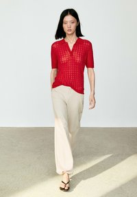 Woman walking wearing a red knit short-sleeve polo shirt, light beige wide-leg pants, and black thong sandals on a plain background.