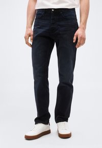 Άνδρας που φοράει σκούρα μπλε τζιν, λευκά αθλητικά παπούτσια με καφέ σόλες και λευκό πουκάμισο, στέκεται μπροστά σε απλό φωτεινό φόντο.