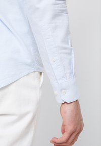 Camisa de manga larga azul clara con puños abotonados, que presenta un tejido texturizado y un dobladillo recto. Los botones son blancos y están firmemente sujetos.
