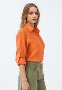 Oranges Button-Up-Hemd aus strukturiertem Stoff, mit hochgerollten Ärmeln und spitzem Kragen. Kombiniert mit hellolivgrünen, hochgeschnittenen Hosen.