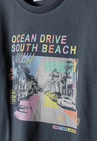 Melnā kokvilnas T-kreklu ar krāsainu grafiku, kas attēlo Ocean Drive, South Beach, ar palmu kokiem un automašīnām. Uz tā ir uzraksts "Lieliskas dienas priekšā."