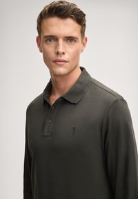 Polo a maniche lunghe verde scuro con placket a due bottoni, colletto e un logo ricamato sul lato sinistro del petto. Tessuto morbido e liscio.
