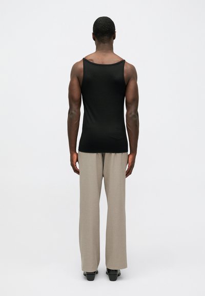 Meesmood riietuses, seistes seljaga, mustas ilma varrukateta särgis, beežides laia säärega pükstes ja mustades kingades, ühtlasel valgel taustal.