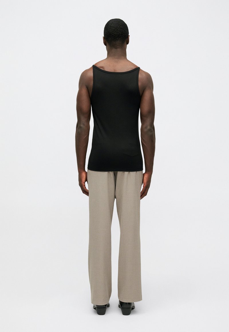 Modello maschile in piedi di spalle, indossando una canotta senza maniche nera, pantaloni beige a gamba larga e scarpe nere su uno sfondo bianco uniforme.