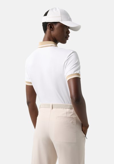 Personne portant un polo blanc avec des bordures beiges, un pantalon beige avec une ceinture tissée, et une casquette de baseball blanche, debout les mains dans les poches.