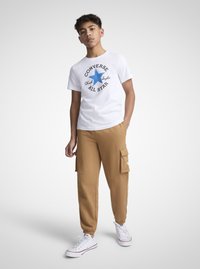 Hvit bomulls t-skjorte med blå stjerne grafikk og svart tekst, kombinert med beige joggebukser med sidelommer og elastiske mansjetter. Hvite joggesko.