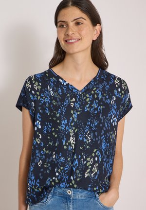 Femme aux cheveux foncés portant un chemisier bleu marine à manches courtes avec un imprimé abstrait bleu, vert et blanc, souriante et posant avec une main dans la poche.