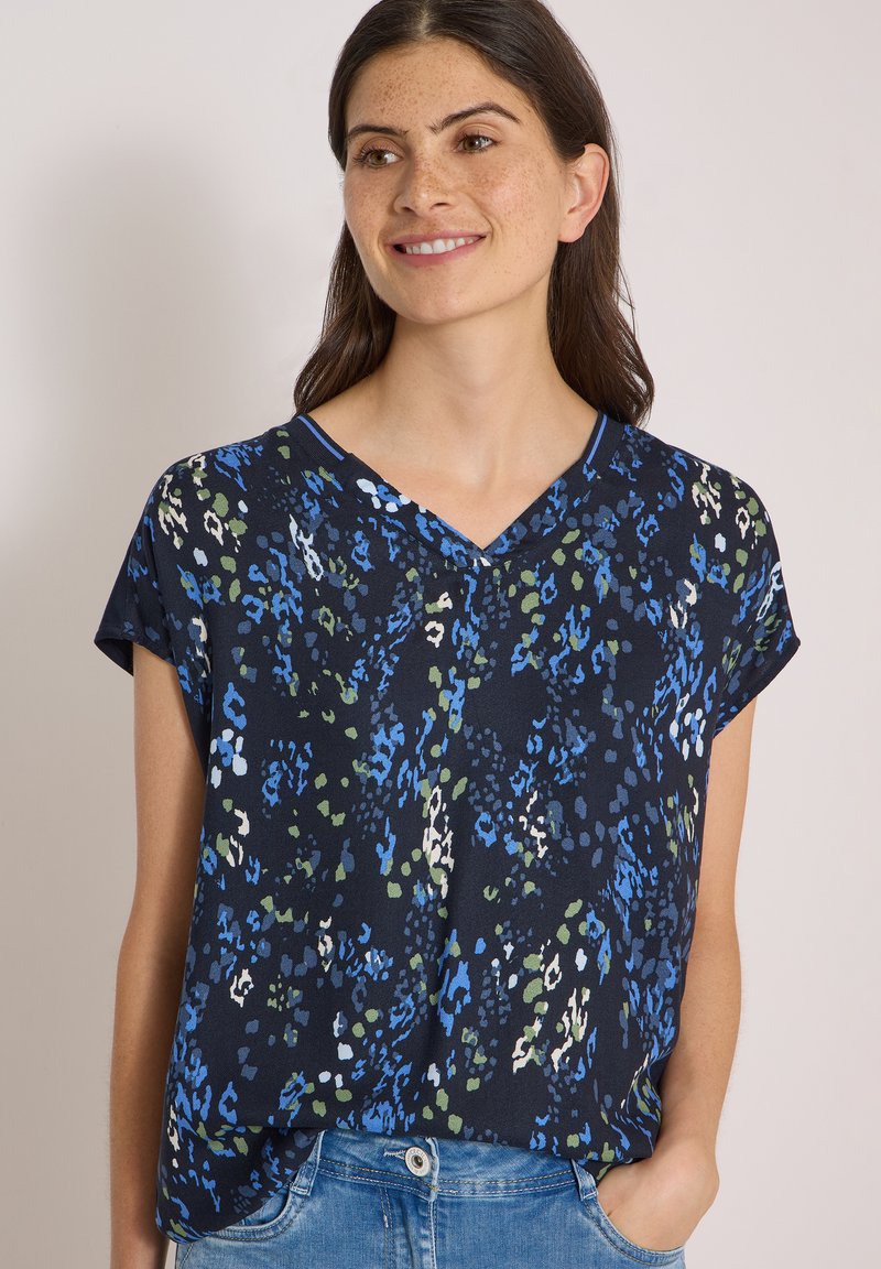 Femme aux cheveux foncés portant un chemisier bleu marine à manches courtes avec un imprimé abstrait bleu, vert et blanc, souriante et posant avec une main dans la poche.
