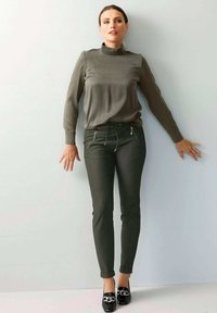 Olivgrünes Langarmoberteil mit hohem Kragen, kombiniert mit schmal geschnittenen, dunkelgrünen Hosen mit Reißverschlusstaschen und schwarzen Mokassins.