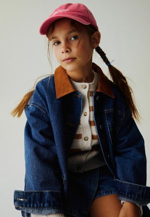 Niña joven con el cabello trenzado, que lleva una gorra rosa, suéter a rayas y una chaqueta vaquera holgada sobre un fondo liso.
