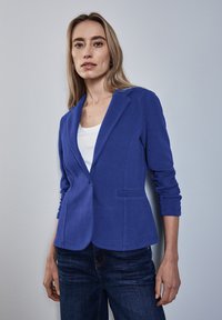 Blå skräddarsydd kavaj med slagkrage, enknappsstängning och två framfickor. Fitted design med långa ärmar och textilstruktur.