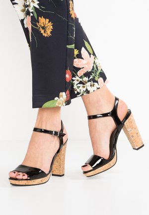 Sorte hæle i laklæder med korkplatform og kraftig hæl. Har en ankelrem med spænde i sølv. Åben tå-design.