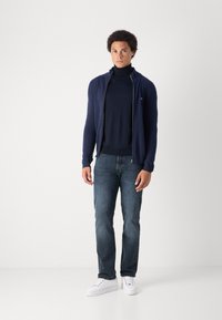 Marinblå zip-up cardigan över en mörk polotröja, ihop med blå jeans och vita sneakers. Mjuk textur, åtsittande design, minimal varumärkesdetalj.