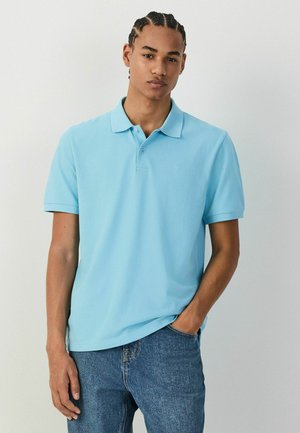 Fiatal férfi cornrow fonatokkal, világoskék galléros pólót és kék farmert visel, egyik kezét a zsebében tartva áll egy egyszerű háttér előtt.