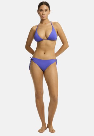 Eine Person steht in einem lila Bikini, die Arme entspannt an den Seiten, mit einem neutralen Ausdruck, vor einem einfarbigen weißen Hintergrund.