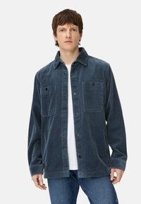 Corduroy overhemdjack in diepblauw, met twee borstzakken, knoopsluiting en een klassieke kraag. Draagt over een wit T-shirt.