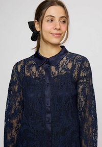 Tengerészkék csipkeblúz hosszú ujjakkal, selyem gallérral, gombos rögzítéssel, virágmintákkal és félig áttetsző anyaggal. Fekete selyem masni a hajban.