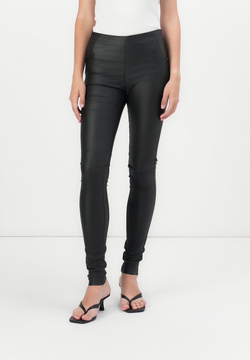Leggings neri in finta pelle con vestibilità slim, vita alta e polsini alla caviglia, abbinati a sandali neri con cinturini. Texture liscia, senza pattern visibili.