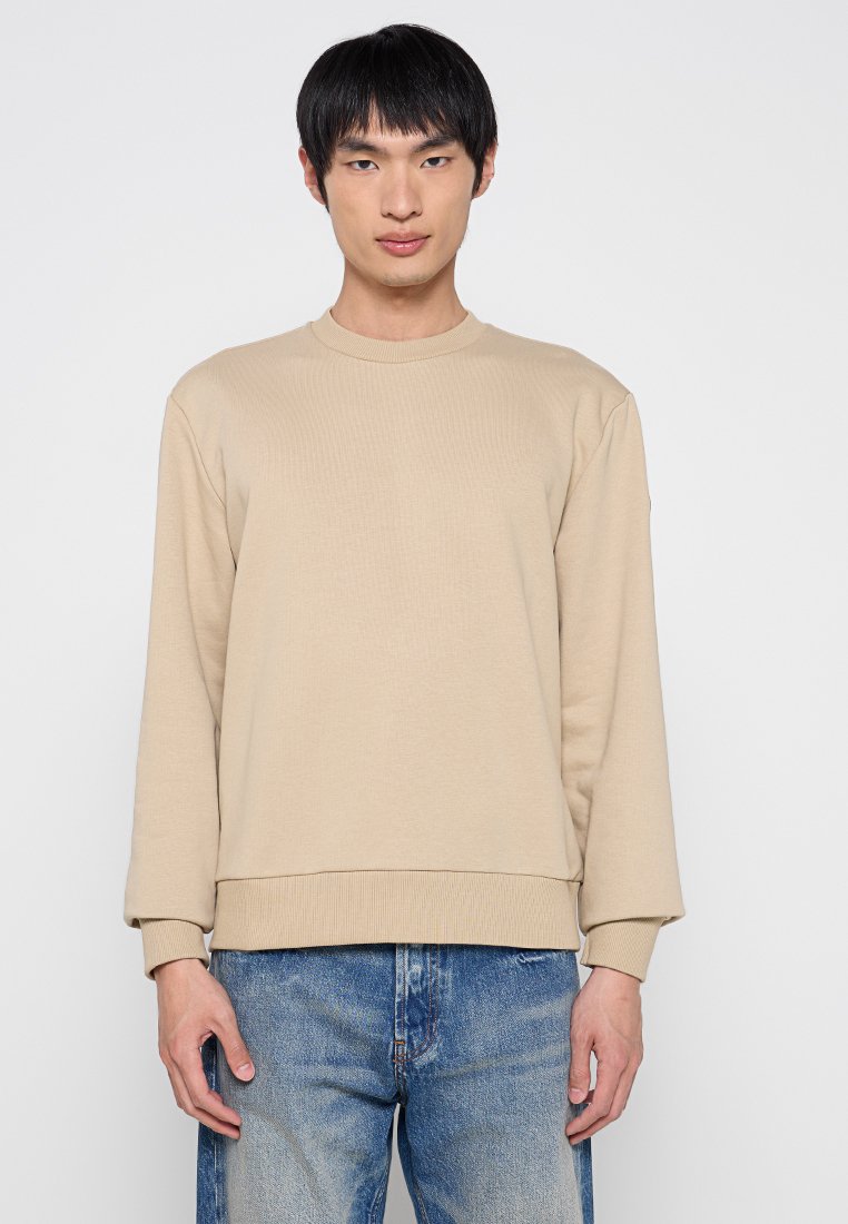 Felpa beige a maniche lunghe con texture a coste, collo rotondo e vestibilità ampia. Abbinata a jeans blu strappati. Design semplice e neutro.