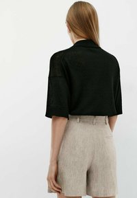 Donna con capelli biondi lisci che indossa una maglietta nera a maniche corte in maglia e pantaloni corti beige in lino a vita alta, mostrata di spalle.