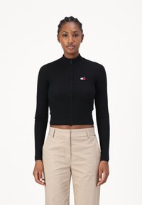 Melns apgrieztais zip-up džemperis ar ribotu tekstūru un garām piedurknēm, ar mazu logotipu sarkanā, baltā un zilā krāsā uz krūtīm.