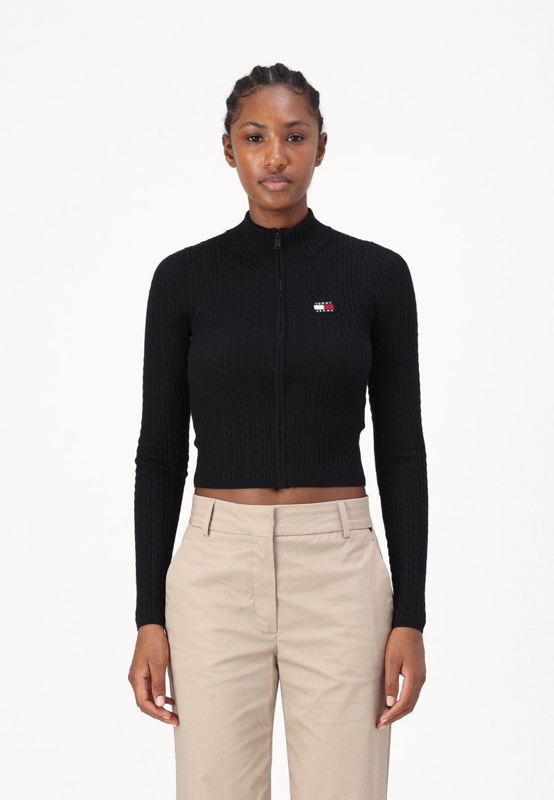 Melns apgrieztais zip-up džemperis ar ribotu tekstūru un garām piedurknēm, ar mazu logotipu sarkanā, baltā un zilā krāsā uz krūtīm.