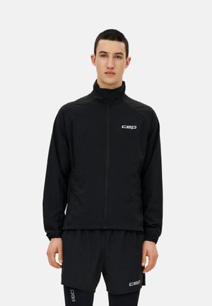 Junger Mann in schwarzer Reißverschlussjacke und passenden Shorts mit weißen "cep"-Logos, stehend vor einem einfarbigen hellgrauen Hintergrund.