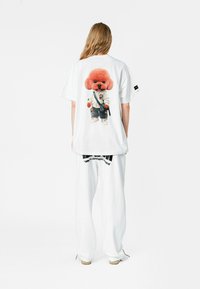 Camiseta blanca oversized con un gráfico colorido de un perro vestido con ropa urbana casual. Modelo combinado con pantalones blancos y zapatos beige.