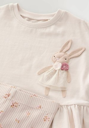 Top in cotone rosa chiaro con applicazione di coniglio, abbinato a un vestito, orlo texturizzato con volant e dettagli floreali sul tessuto inferiore.