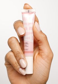 Blush en suero rosa en un tubo suave, con una punta texturizada y etiqueta de texto. Sujeto por una mano con uñas lisas en tono nude sobre un fondo blanco.