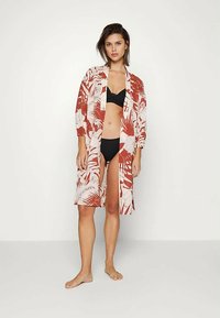 Femeie în picioarele goale, purtând un bikini negru și un halat deschis cu imprimeu tropical în nuanțe de roșu cărămiziu și alb, în fața unui fundal alb simplu.