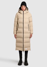Manteau long beige rembourré avec une fermeture éclair, deux poches latérales et une capuche noire. Associé à des leggings noirs et des bottes noires épaisses.