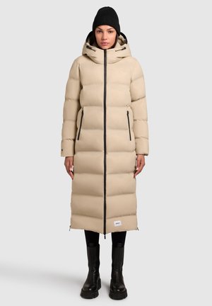 Beige gewatteerde lange jas met een rits, twee zijzakken en een zwarte capuchon. Gecombineerd met zwarte leggings en robuuste zwarte laarzen.