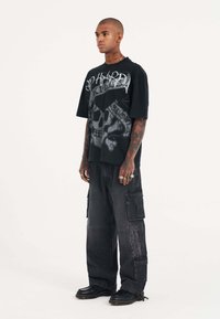 Camiseta gráfica negra con diseño de calavera, combinada con pantalones cargo negros holgados. Los detalles incluyen múltiples bolsillos y un acabado texturizado.