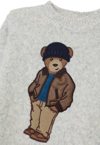 Gestickter Teddybär mit einer marineblauen Mütze, braunem Lederjacke, blauer Sweatshirt, beigen Hosen und Schuhen auf einem hellgrauen Strickstoff.