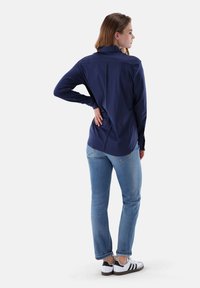 Donna in piedi, che indossa una maglietta a maniche lunghe blu navy, jeans azzurri e sneakers bianche, con una mano appoggiata sulla parte bassa della schiena.