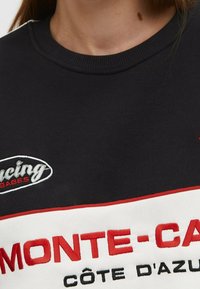 Zwarte sweatshirt met wit en rood gestreept ontwerp. Voorzien van geborduurde tekst "MONTE-CARLO" en "CÔTE D'AZUR" in rood en zwart. Geribbelde halslijn.