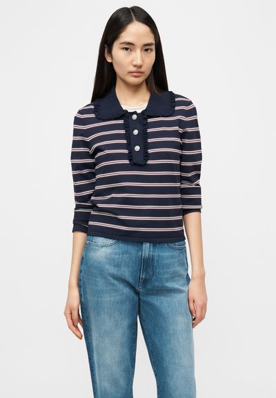 Μπλούζα με ναυτικό μπλε και λευκές ρίγες, με βολάν στο γιακά, τρία κουμπιά στον λαιμό και μανίκια τριών τετάρτων, συνδυασμένη με ανοιχτό μπλε τζιν.