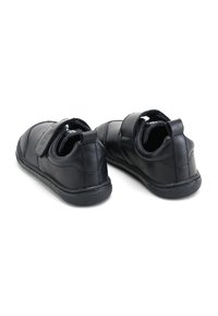 Zapatos de cuero negro con un perfil bajo, que cuentan con una correa de velcro y una suela de goma texturizada, diseñados para la comodidad y la facilidad de uso.