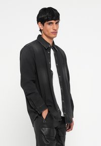 Chemise en denim noir avec un devant à boutons, des manches longues et un col pointu, superposée à un t-shirt blanc, modèle portant un pantalon noir.