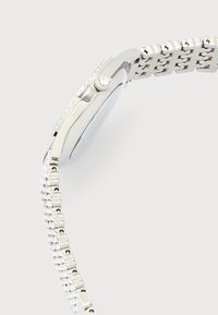 Ασημένιο ρολόι χειρός με μπρασελέ από μεταλλικούς κρίκους και κορώνα με αυλακώσεις που φέρει ένα μικρό διαφανές λίθο, εμφανιζόμενο από το πλάι σε ανοιχτό φόντο.