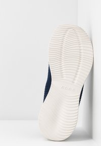 Semelle d'une chaussure navy bleue à enfiler avec une semelle en caoutchouc texturée blanche montrant un motif de bande de roulement ondulée et la marque "BOBS".