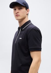 Μαύρο πόλο με λευκές λεπτομέρειες, με φερμουάρ μέχρι τη μέση, κοντά μανίκια και λογότυπο BOSS στο στήθος. Φοριέται με σκούρο καπέλο.